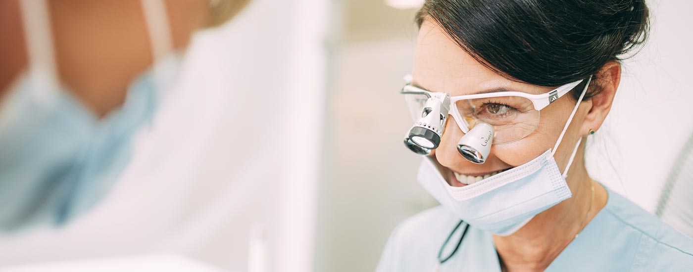 Zahnärztin Heidi Prutean trägt während einer Wurzelbehandlung eine Lupenbrille und lacht ihre Assistentin an.